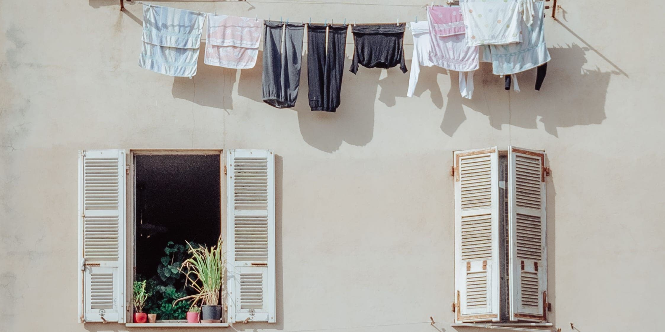 Washing hung on a line across a sunlit plaster facade, between two shuttered windows