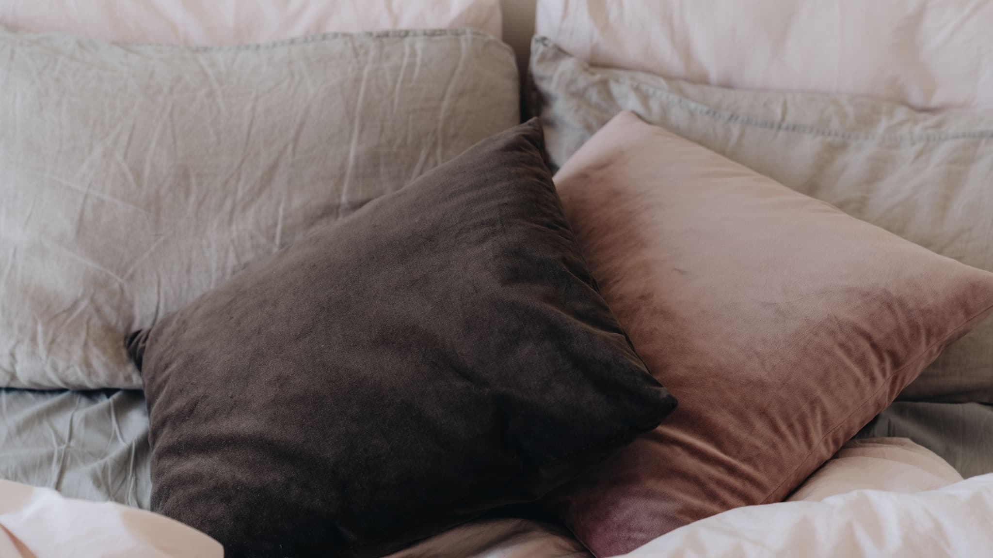 Pillows piled against a headboard, pale pink cases and one in sage linen, with a brown and a dusty-pink velvet cushion in front
