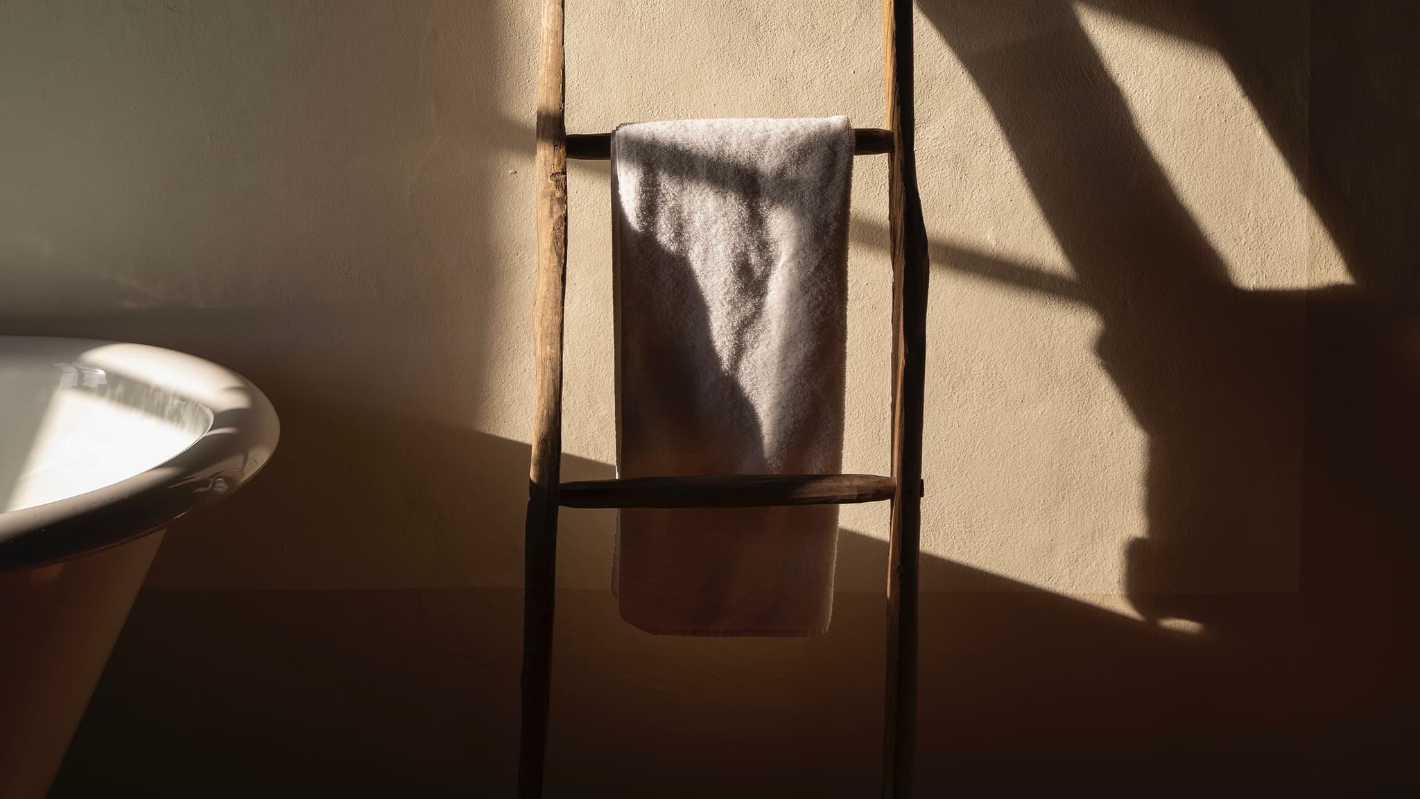 A pale towel folded over the rung of a wooden ladder against a plaster wall, low sun raking across it, the rim of a bath at the left