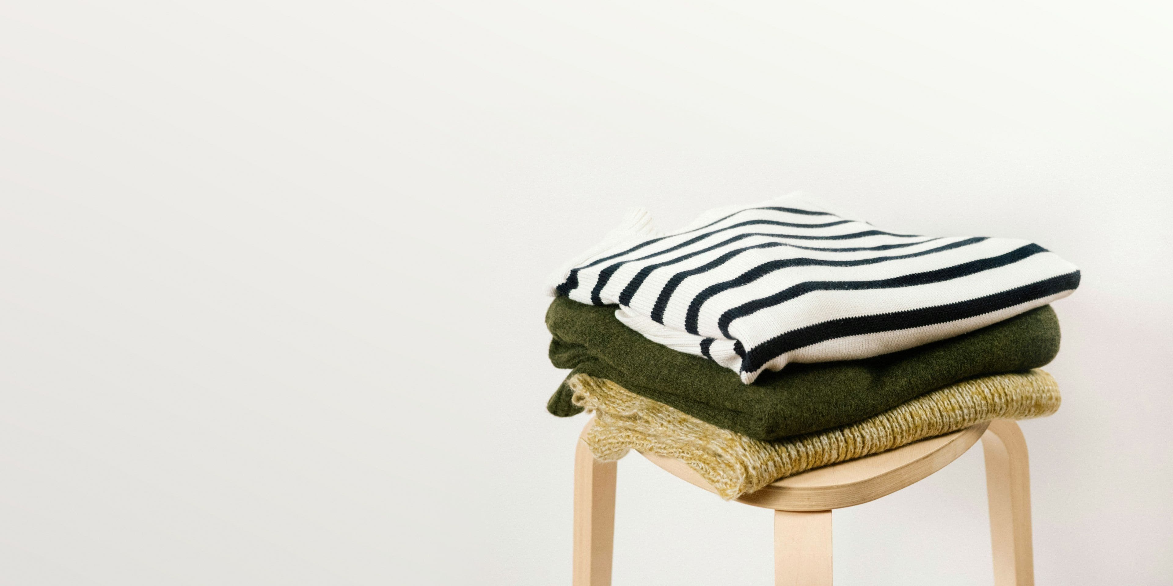Three folded jumpers stacked on a pale wooden stool against a plain wall: a cream one with navy stripes on top, then olive, then a speckled yellow knit