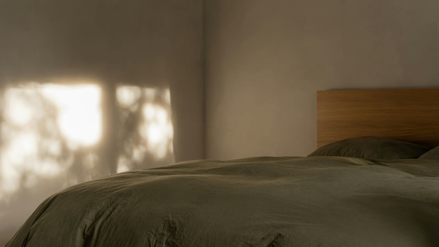 A bed made up in olive linen against a warm plaster wall, late sun throwing the shadows of leaves across the wall at the left
