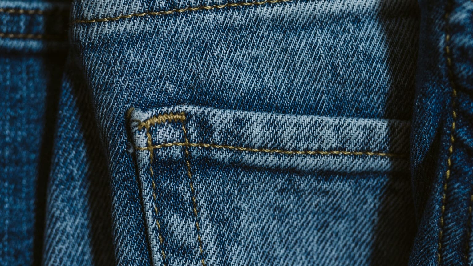 The back of a pair of mid-blue jeans, close: the corner of a back pocket with its bar-tack, the yoke seam above, darker denim either side