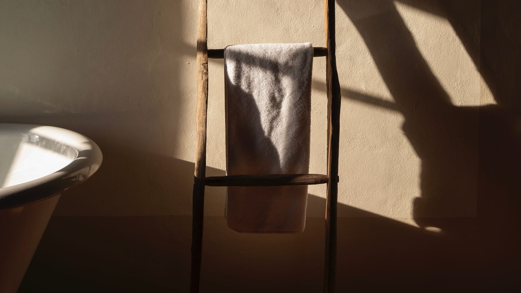 A pale towel folded over the rung of a wooden ladder against a plaster wall, low sun raking across it, the rim of a bath at the left