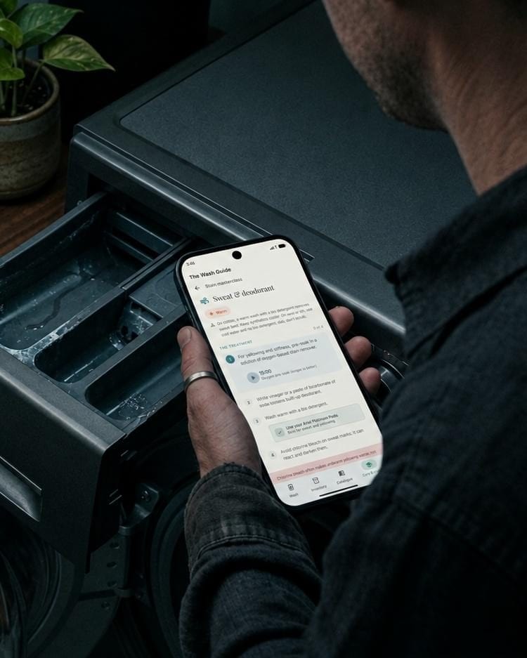 A hand holding a phone showing The Wash Guide at the open detergent drawer of a washing machine, in low evening light