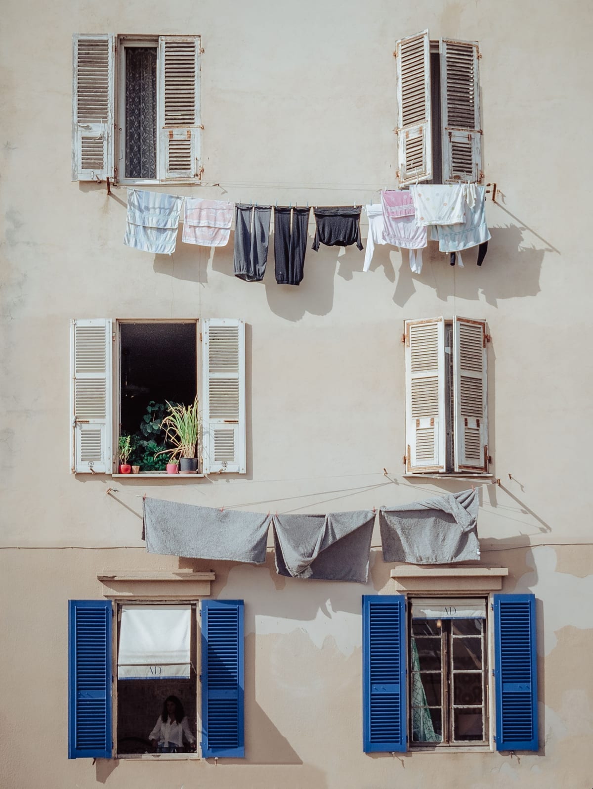 A sunlit plaster facade with washing hung on a line between two shuttered windows, a plant on the sill below