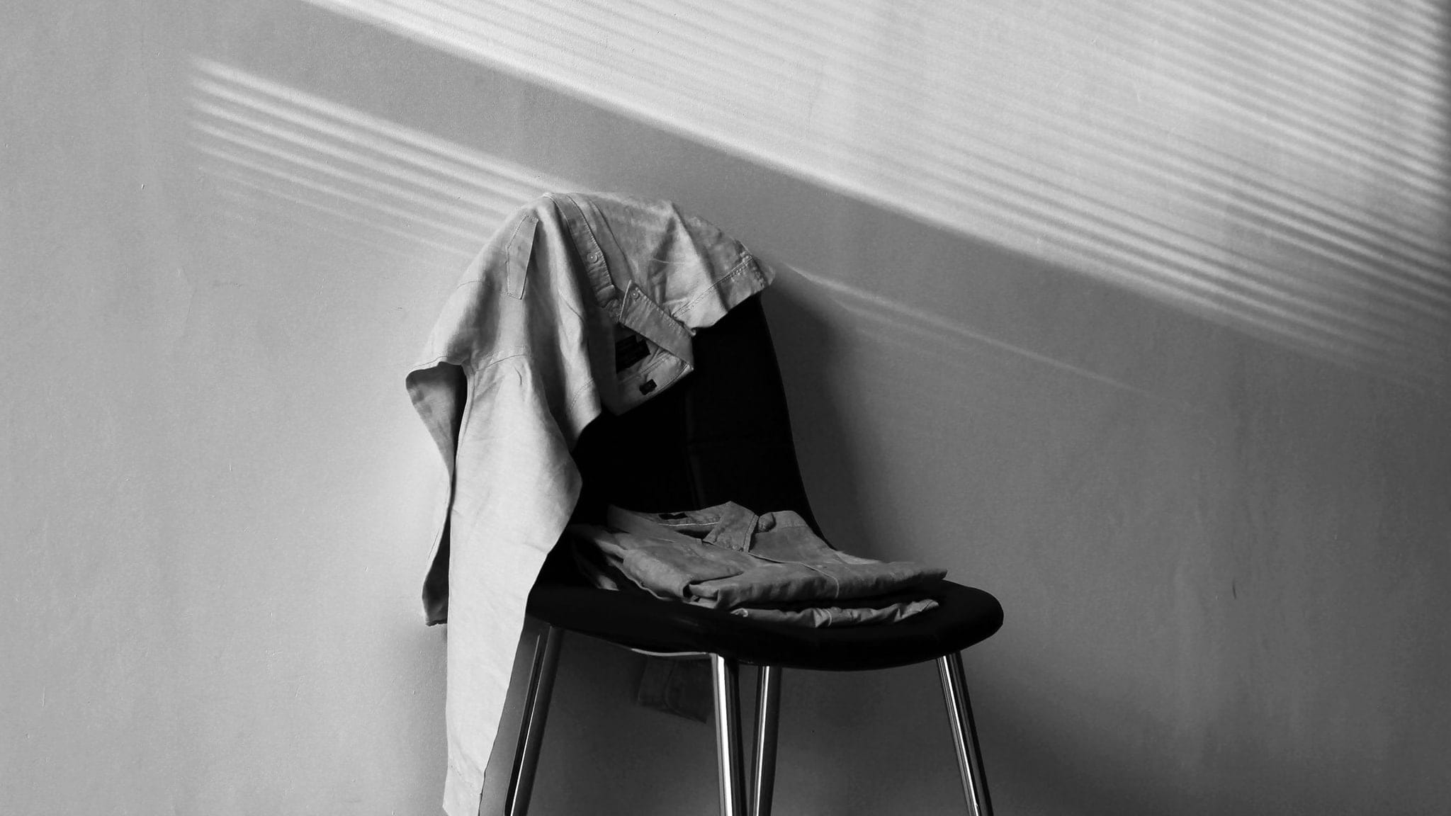 A shirt hung over the back of a chair with jeans and a folded tee on the seat, blind-light striping the wall behind