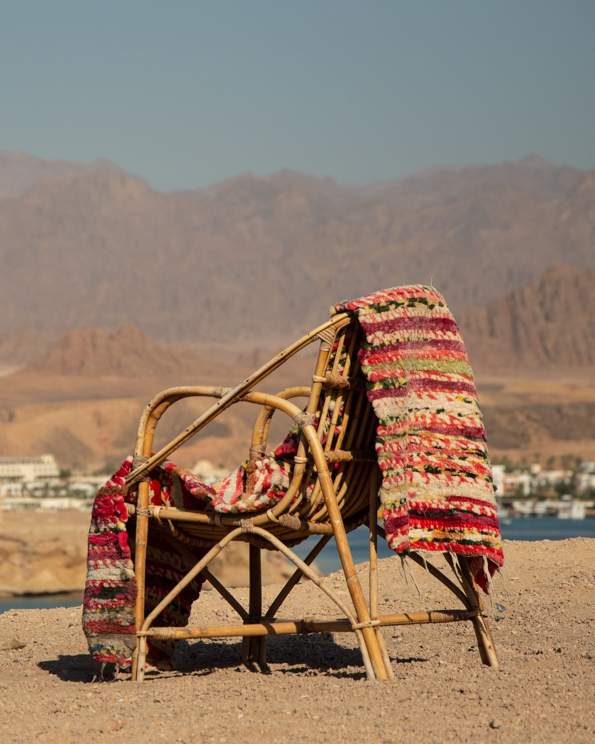 A cane chair on a bare, sunlit shore with a bright woven rug aired over its back, blue water and desert mountains behind it